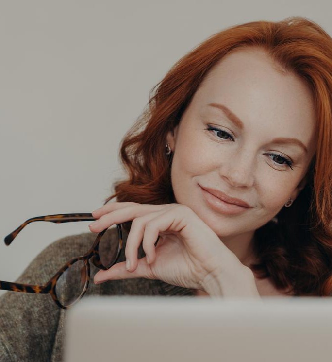Photo of redhead woman takes off glasses, enjoys free time for e learning, focused at monitor of laptop computer, does distance job. Female student prepares for exams, uses modern technologies